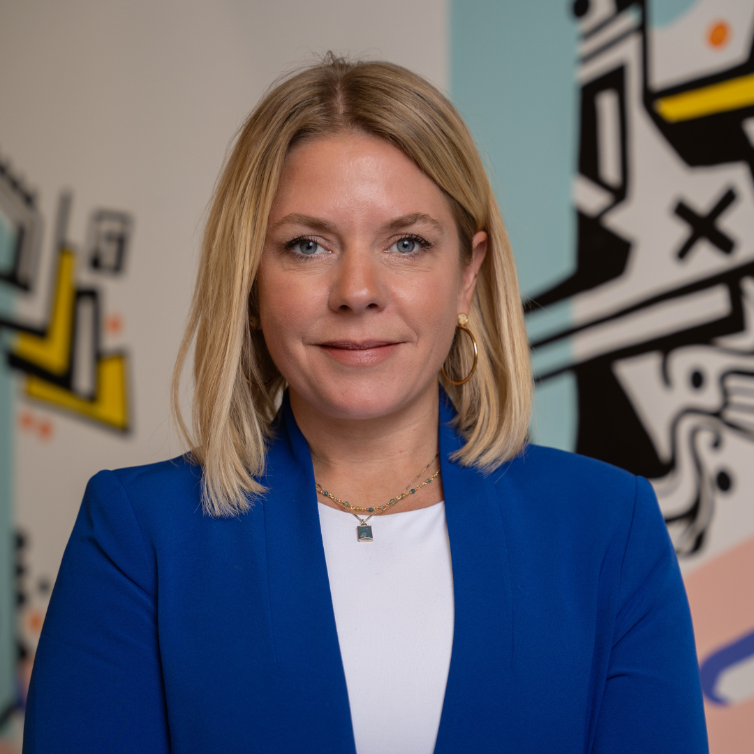 Amy Peterson, founder of Rebel Nell with shoulder-length blonde hair wearing a royal blue blazer and white blouse stands in front of a colorful mural background, smiling slightly at the camera.
