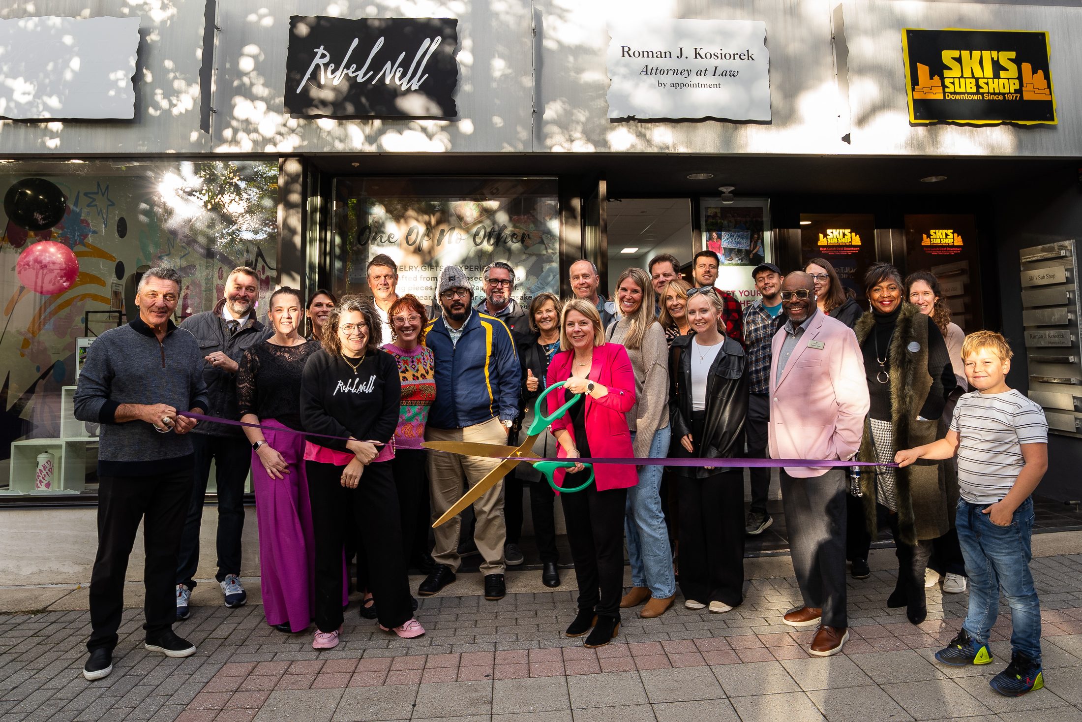 A large group of people stands outside the new Rebel Nell storefront for a ribbon-cutting ceremony. The group includes staff, community members, and supporters, with Amy Peterson in a bright pink blazer holding oversized scissors at the center.