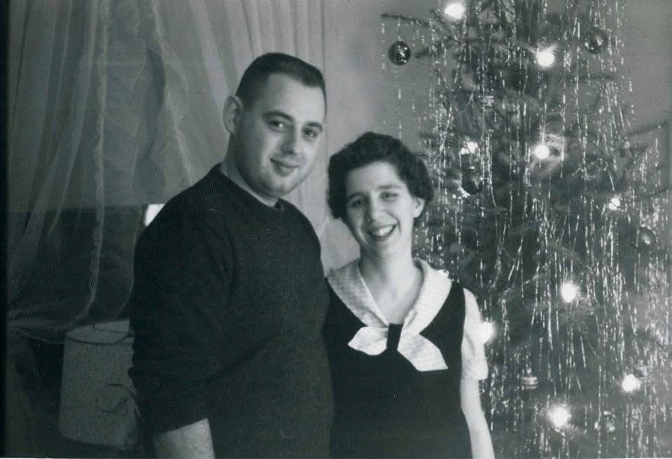 A black and white vintage photograph of a young man and woman smiling together in front of a decorated Christmas tree with tinsel and lights, likely taken in the 1950s or early 1960s.
