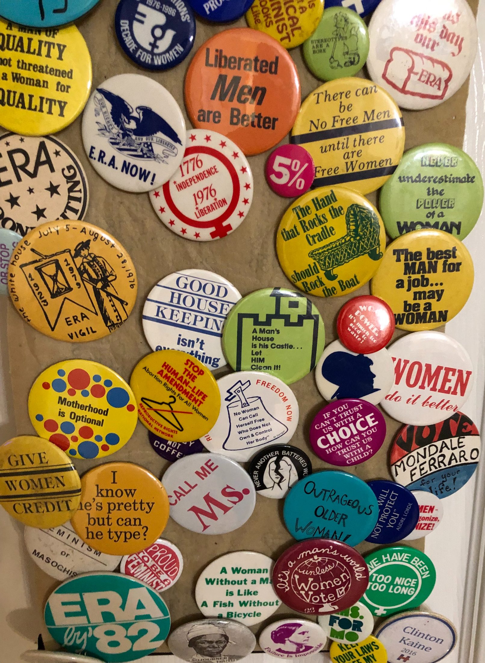 A colorful collection of vintage feminist and women's rights campaign buttons from the 1970s–1980s, featuring slogans such as 'E.R.A. Now,' 'Liberated Men are Better,' 'Motherhood is Optional,' 'Call Me Ms.,' 'Women Do It Better,' and 'ERA by '82,' among many others, displayed on a cork board.