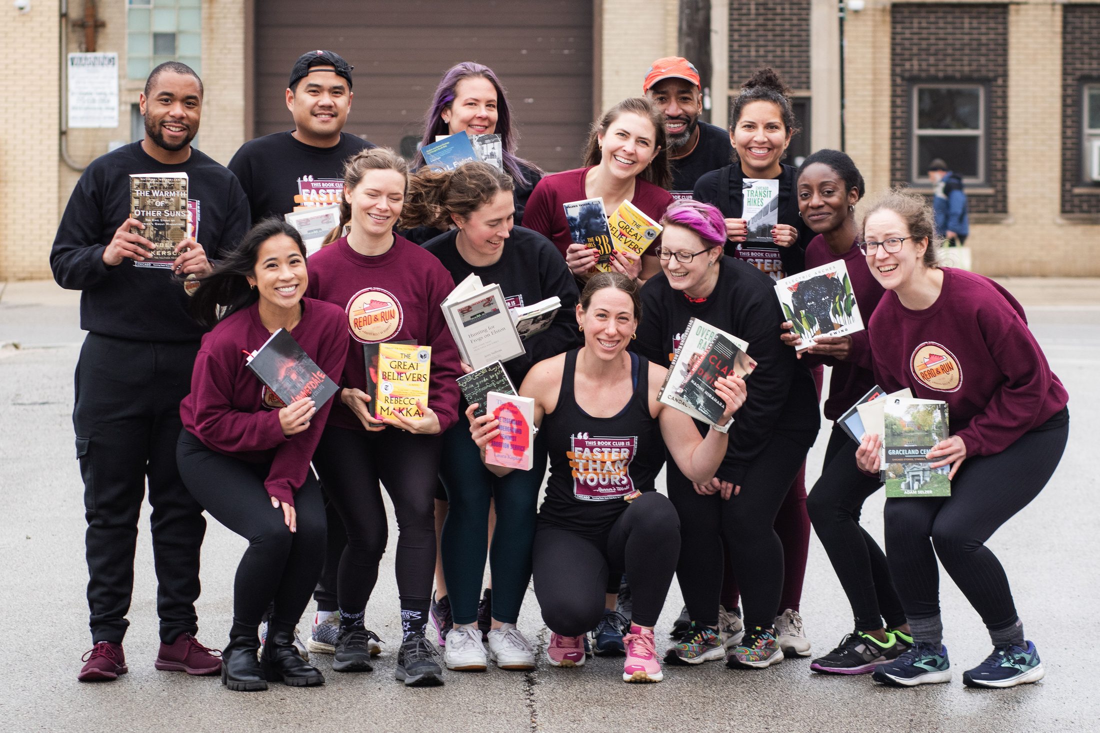 A diverse group of about a dozen smiling adults pose together outdoors on a city street, holding up various books. Many are wearing maroon “Read & Run” shirts or sweatshirts, suggesting they are part of a literary running club. The group stands closely together, creating a warm, community-focused atmosphere that blends fitness and reading.