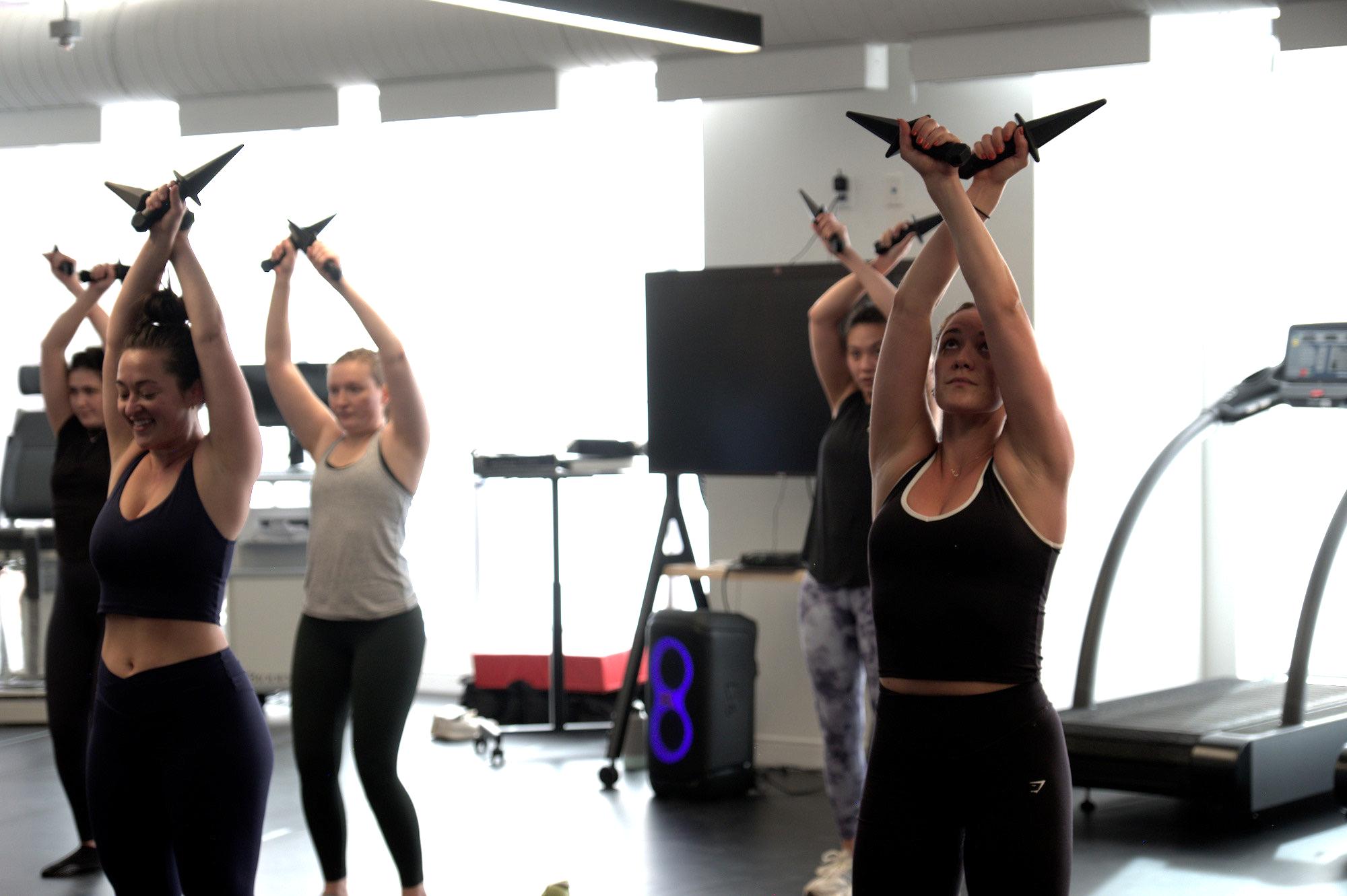 Four women exercise inside a bright fitness studio, lifting dagger-shaped dumbbells overhead in unison. They wear athletic clothing and stand near treadmills and a speaker, with natural light streaming through large windows behind them. The scene suggests a themed group workout class focused on strength training.