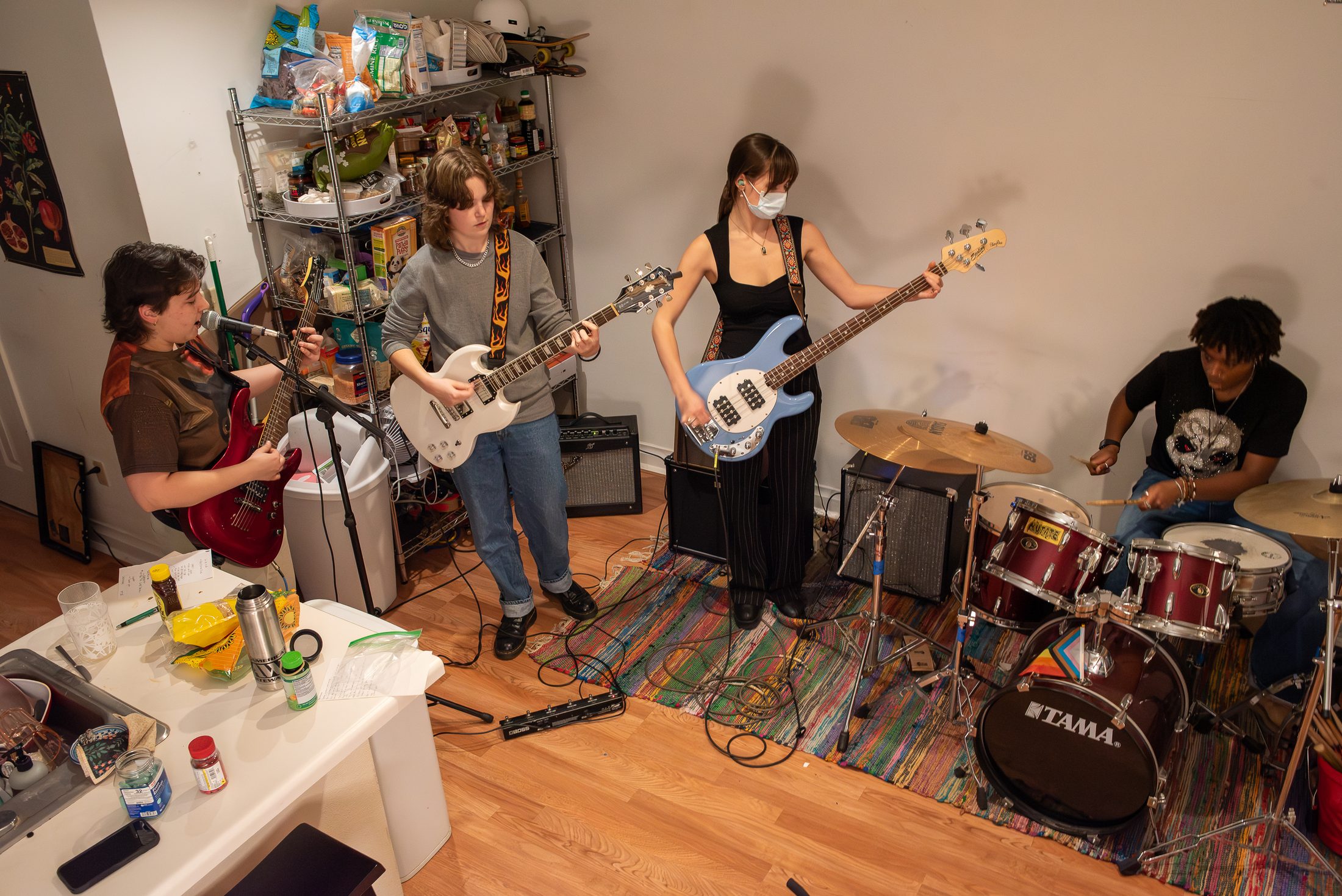 Four band members rehearse in a small apartment kitchen and living room. Three play electric guitars and bass while one plays a drum kit set up on a colorful rug. Cables run across the wooden floor, and a metal shelving unit stocked with snacks and household items stands behind them, emphasizing the intimate, DIY setting.