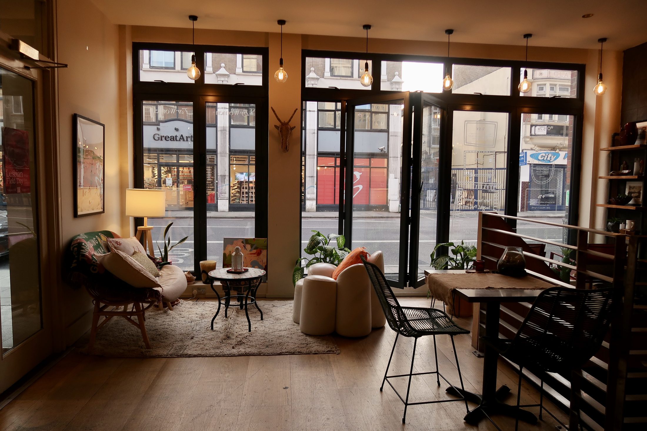A warm, inviting café or lounge interior with large floor-to-ceiling windows looking out onto a city street. The space features wooden floors, a cozy seating area with a cushioned chair and sofa, a small round table with a lamp, and bar-height seating along the right wall. Natural light fills the room, giving it an intimate, neighborhood gathering spot feel.