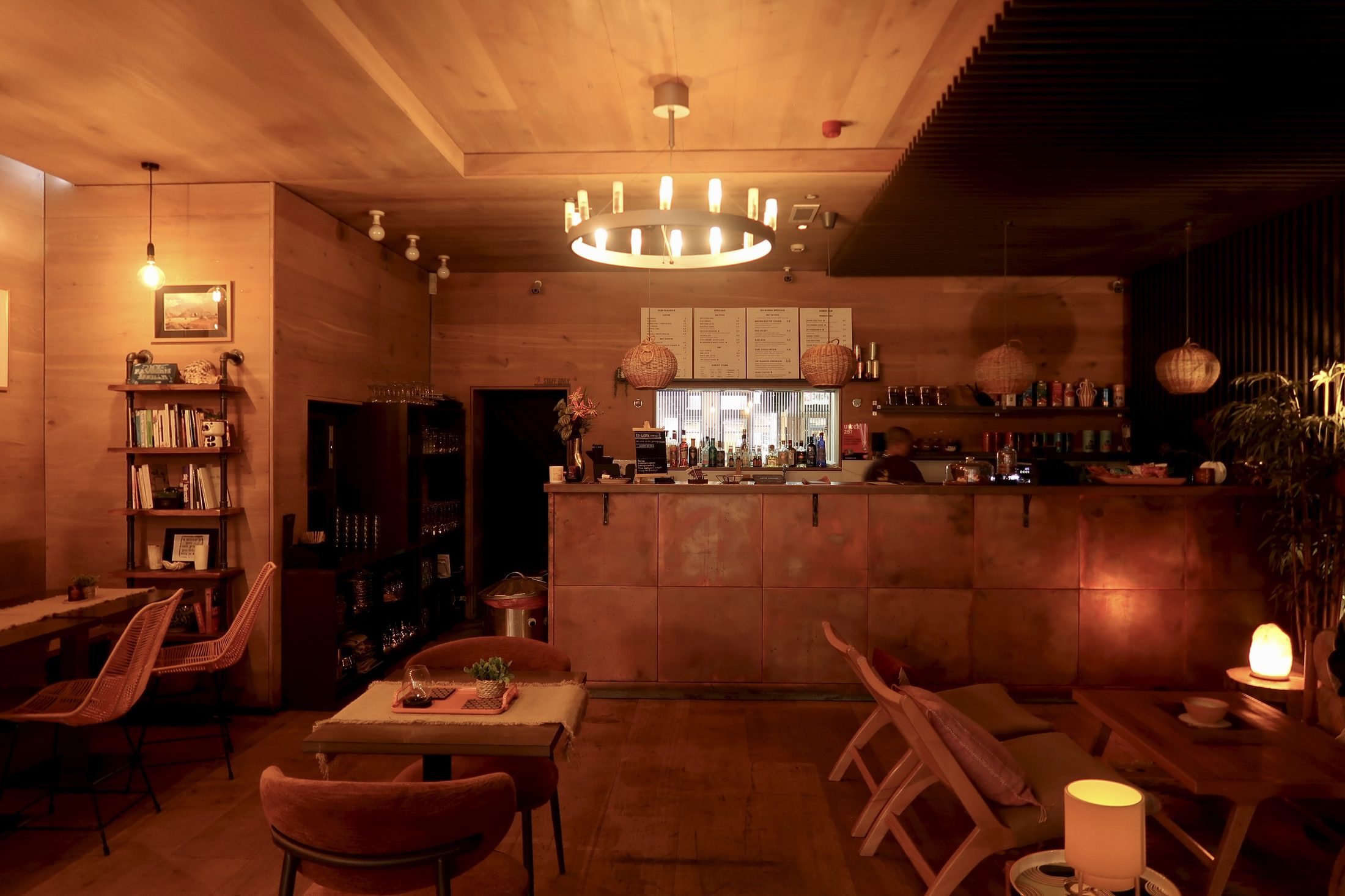 The warm, dimly lit interior of a bar or café at night. A circular chandelier with exposed Edison bulbs hangs above a wooden bar counter lined with bottles and menus. Velvet chairs surround low tables in the foreground, a bookshelf lines the left wall, and wicker pendant lights hang above the bar. Candles and a Himalayan salt lamp cast a soft amber glow throughout the intimate space.