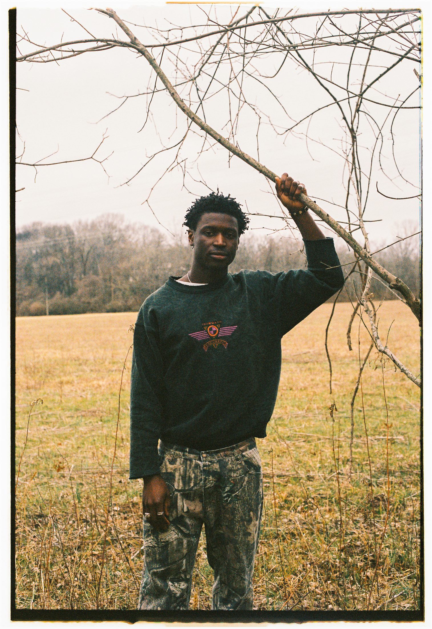 A Black man stands in an open field in winter, holding a bare tree branch overhead. He wears a dark sweatshirt and patterned pants, looking calmly toward the camera as leafless trees and muted grass stretch into the background.