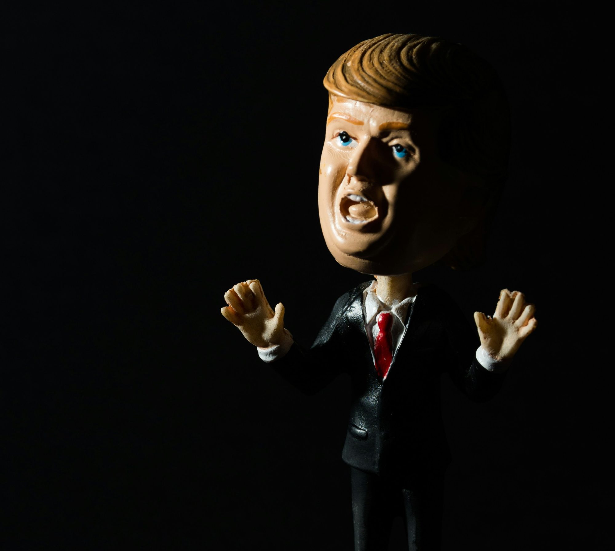 Caricature figurine of a shouting male political figure, partially illuminated from the side, surrounded by darkness.