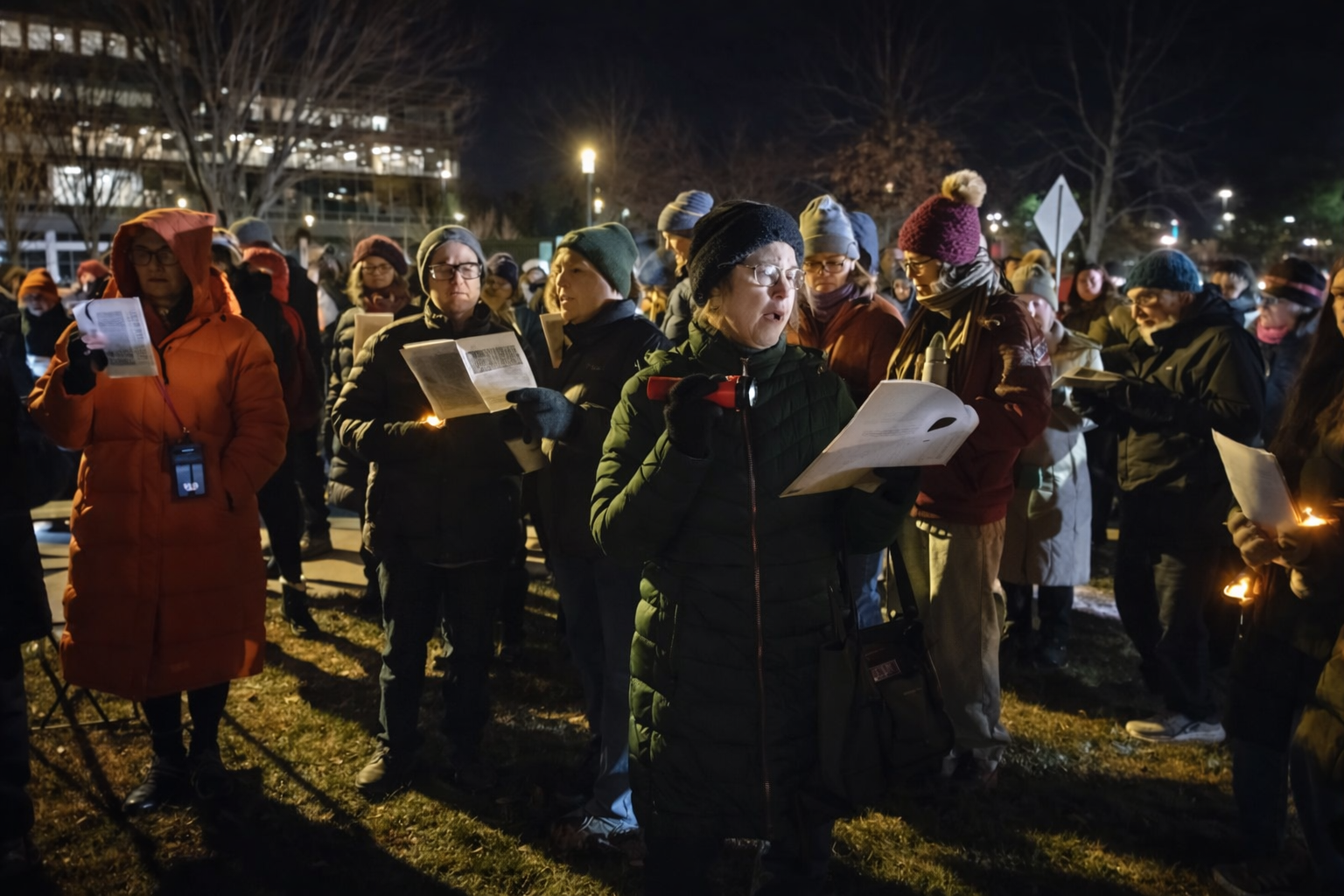 A group of bundled-up people stand outdoors at night holding candles and printed pages, appearing to sing or read together during a vigil. Streetlights and bare winter trees are visible in the background.