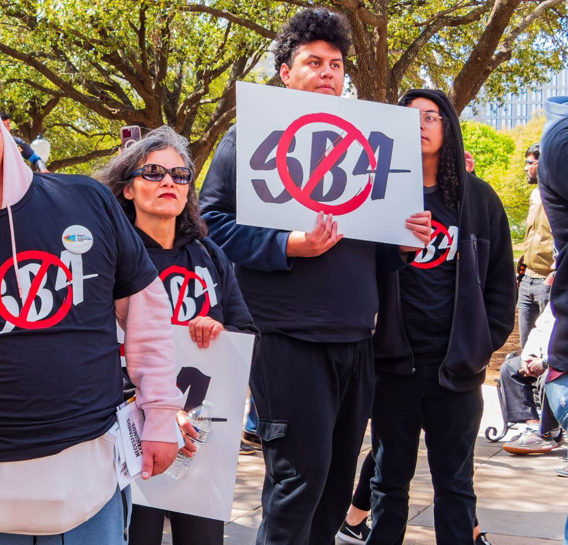 A group of protesters stand outdoors holding signs opposing Texas Senate Bill 4. One person holds a sign reading “SB4” crossed out with a red prohibition symbol, while others wear black shirts with similar anti-SB4 symbols. Trees and additional demonstrators are visible in the background.