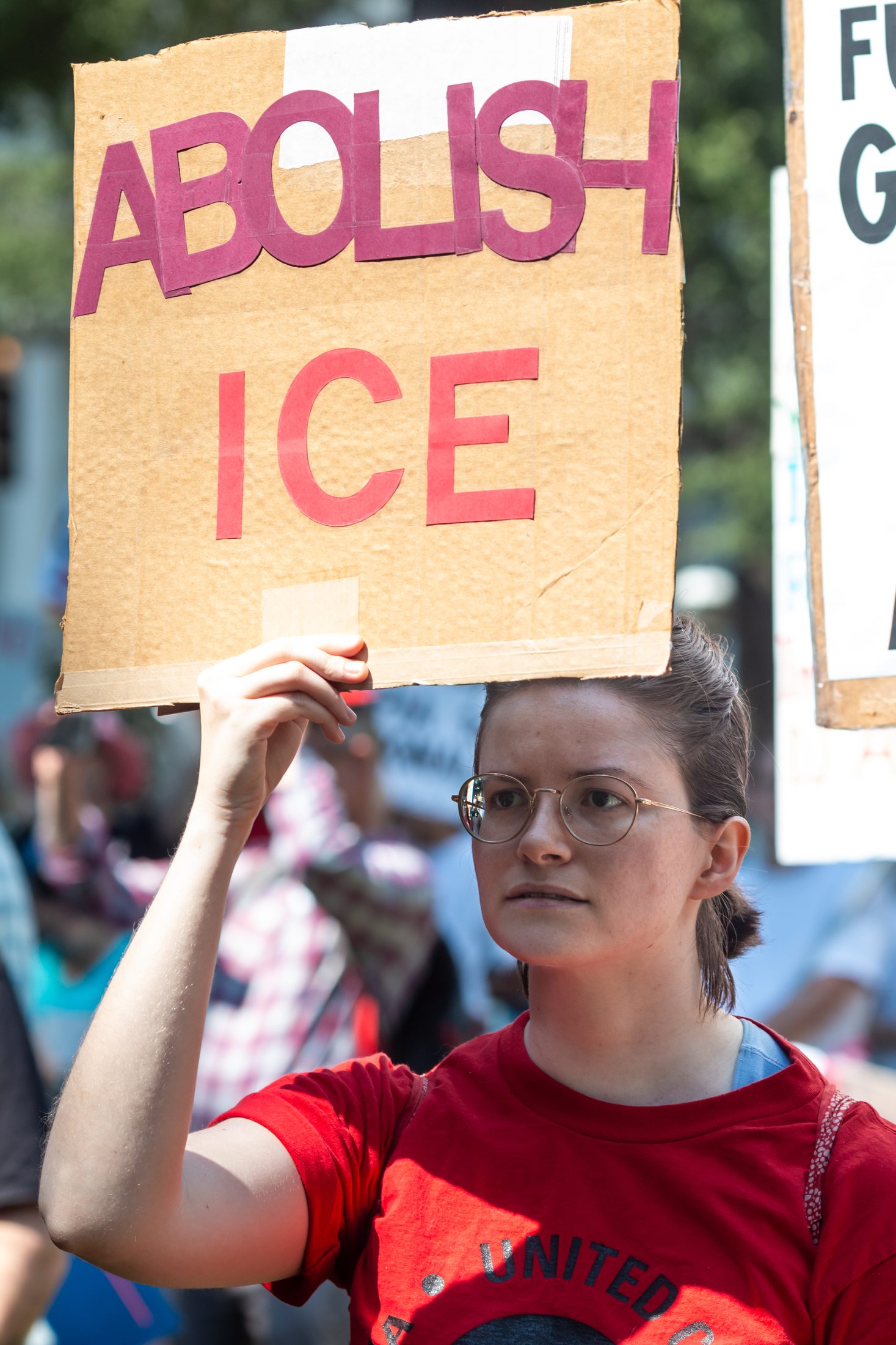 A woman wearing glasses and a red shirt holds a cardboard protest sign reading “Abolish ICE” above their head during a demonstration. Other protesters and signs are visible blurred in the background.