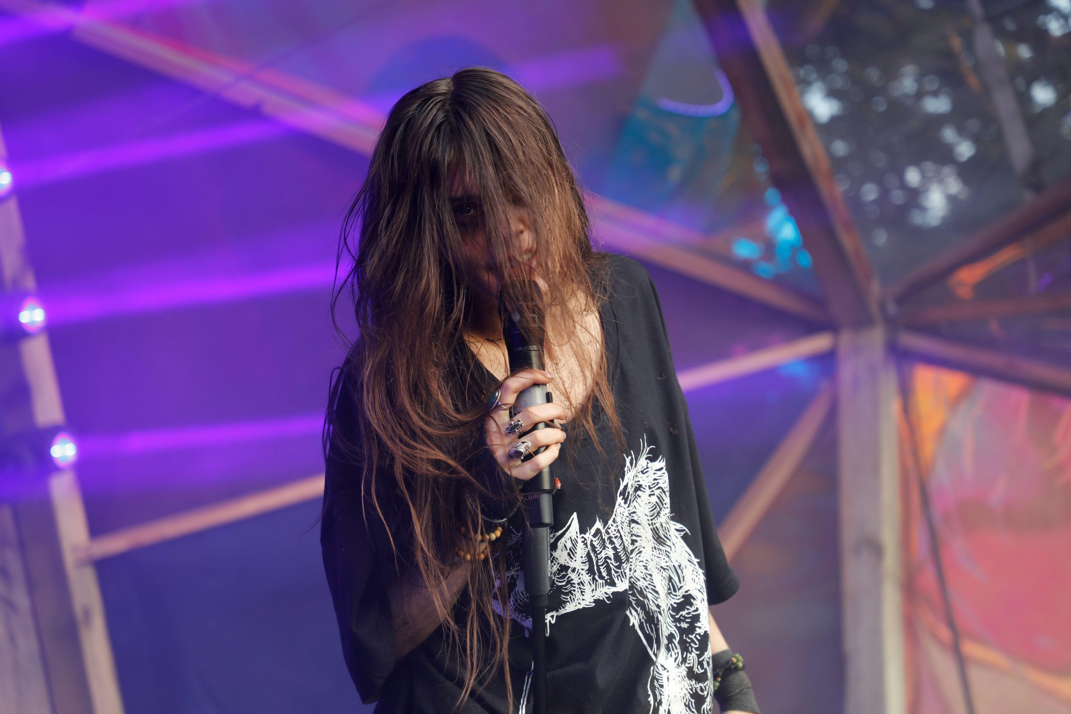 11:19Claude responded: A performer with long dark hair falling across their face holds a microphone on stage, wearing an oversized black graphic t-shirt and rings on their fingers, b…A performer with long dark hair falling across their face holds a microphone on stage, wearing an oversized black graphic t-shirt and rings on their fingers, bathed in purple stage lighting.