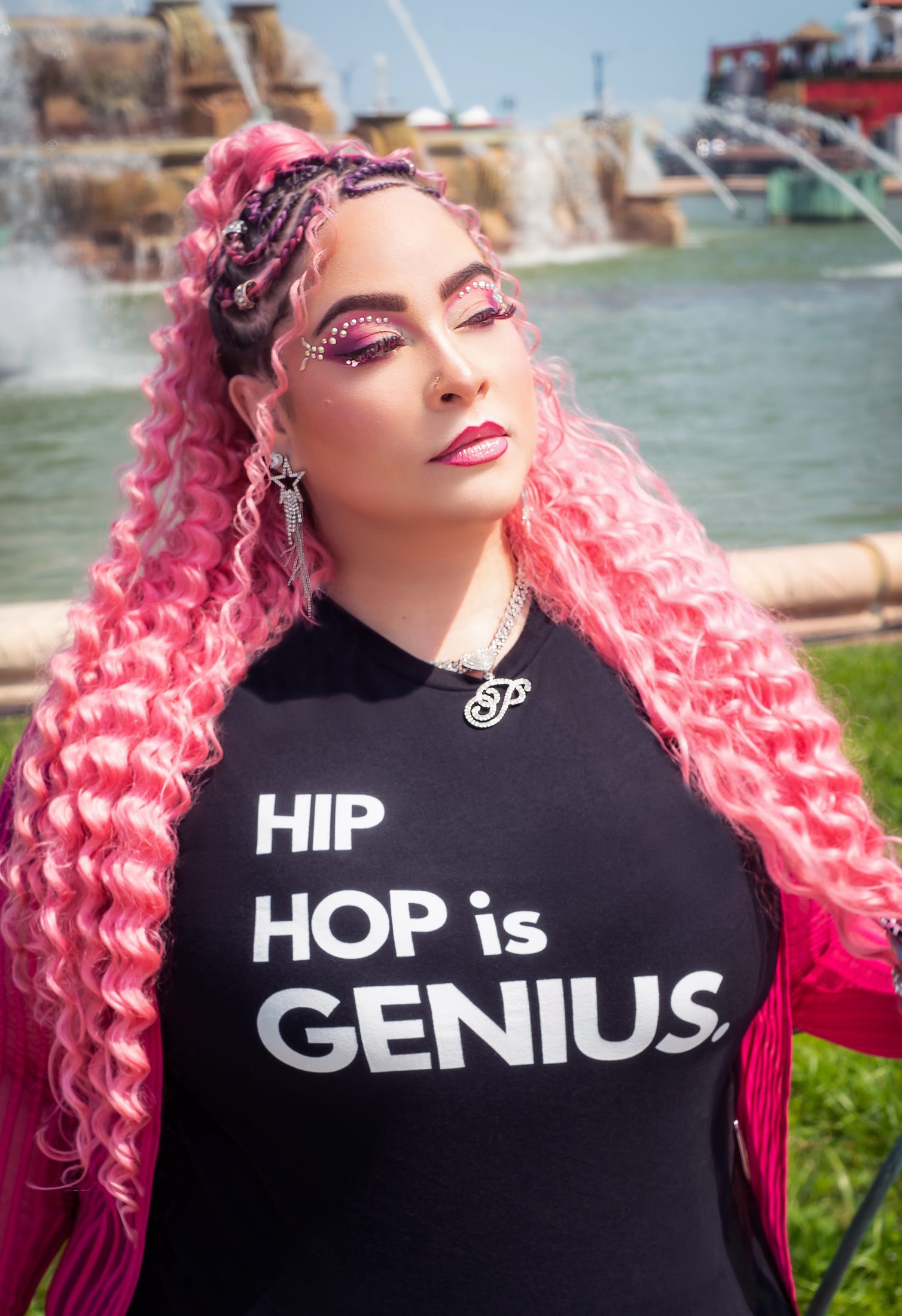 Pinqy Ring stands outdoors in front of a fountain, wearing a black T-shirt reading “Hip Hop is Genius,” with long pink curly hair, braided at the crown, and rhinestone makeup around her eyes.