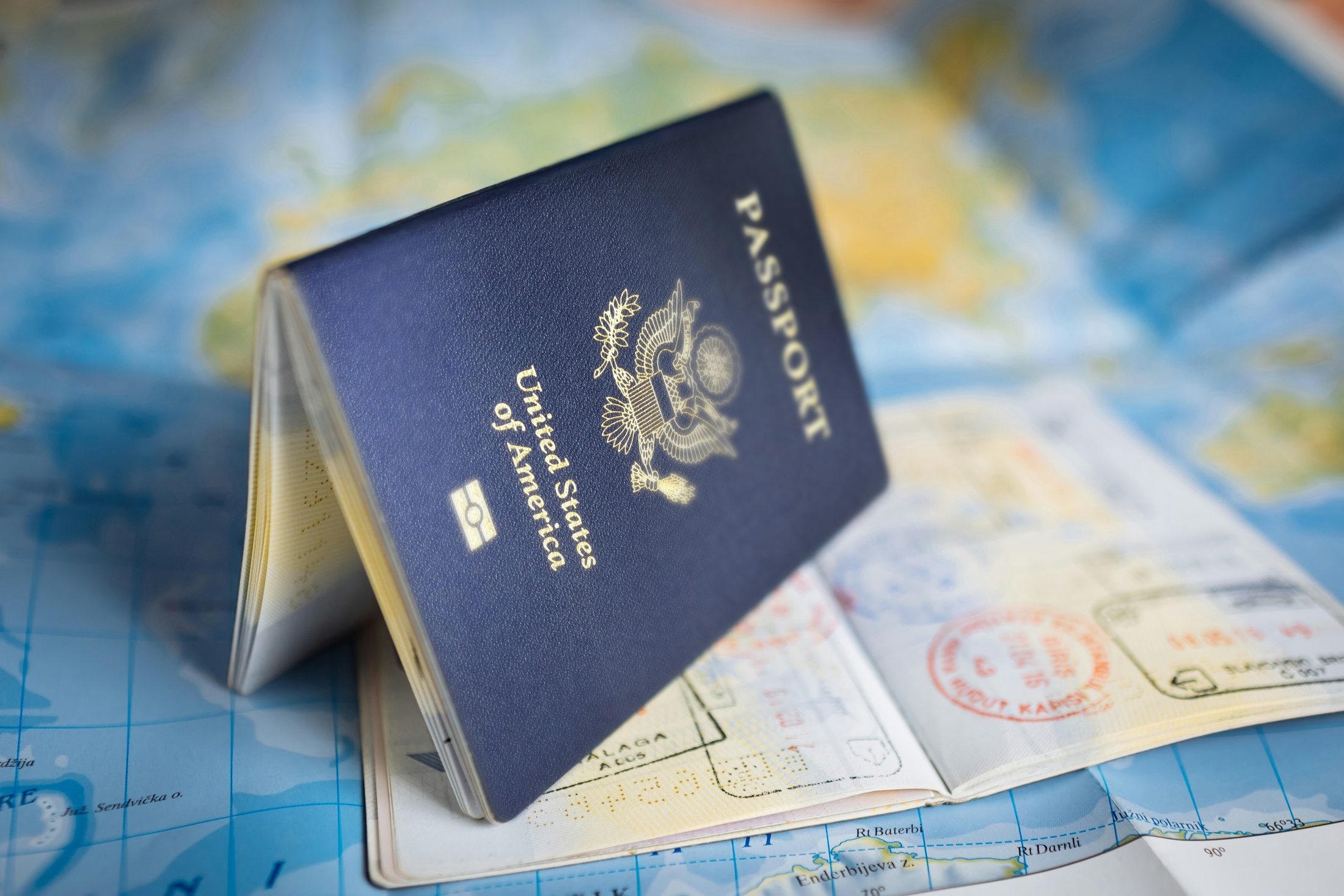A blue United States passport standing slightly open on top of an unfolded world map, with visible entry stamps inside, suggesting international travel and mobility.