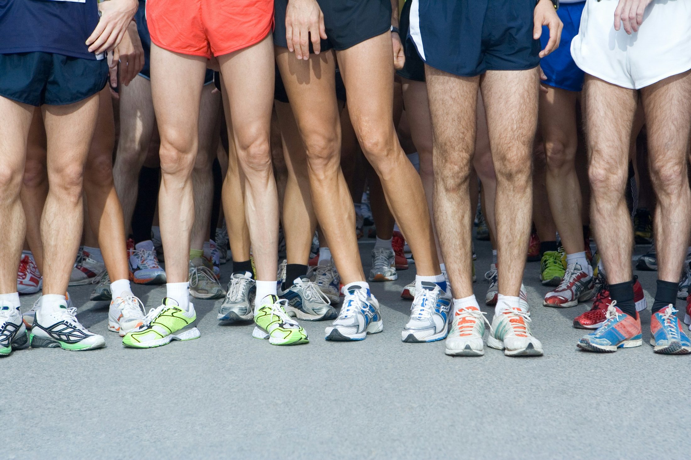 A group of runners stands shoulder to shoulder at a starting line, shown from the waist down, wearing shorts and colorful running shoes on a paved road.