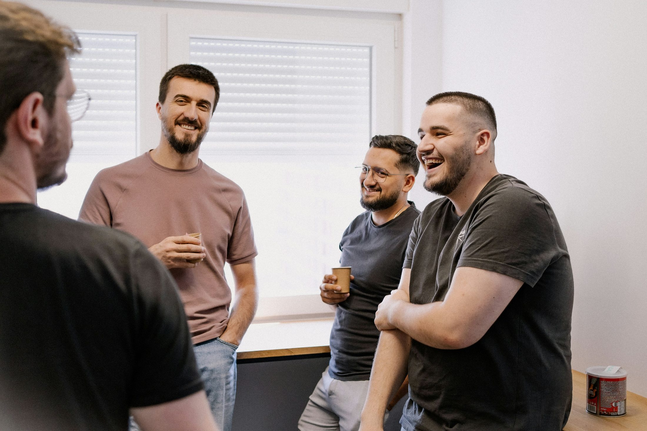 Four men stand together in a bright room, laughing and talking casually. Two are holding small paper cups. The mood is relaxed and candid, suggesting an informal group conversation or discussion circle.