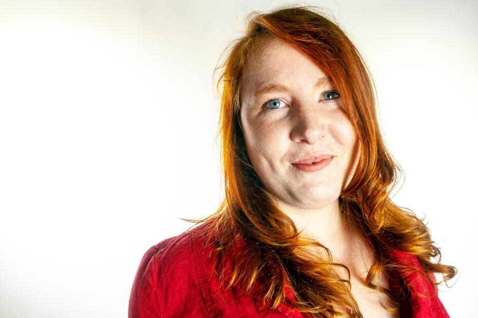 Portrait of Sophie Lewis. She has long, wavy red hair and blue eyes, and is wearing a red shirt. She is photographed against a white background, looking slightly upward with a soft expression.