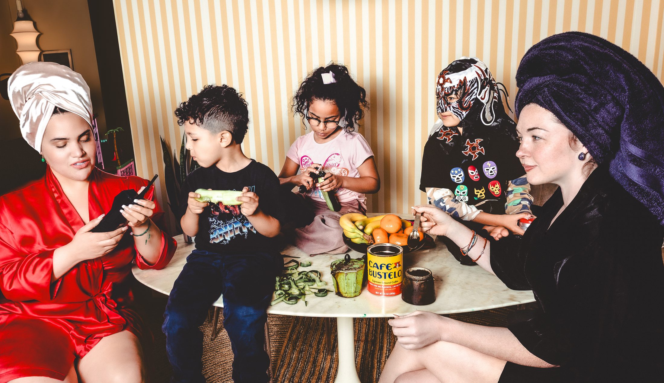 Two women and three children sit together around a small round table covered with fruit, vegetables, and a can of Café Bustelo. The women wear elaborate head wraps and robes — one in red satin, one in black velvet — while the children eat and play. Two of the children wear novelty wrestling masks. The scene is warm and intimate, set against a yellow striped wallpaper background, evoking a sense of playful domestic community.