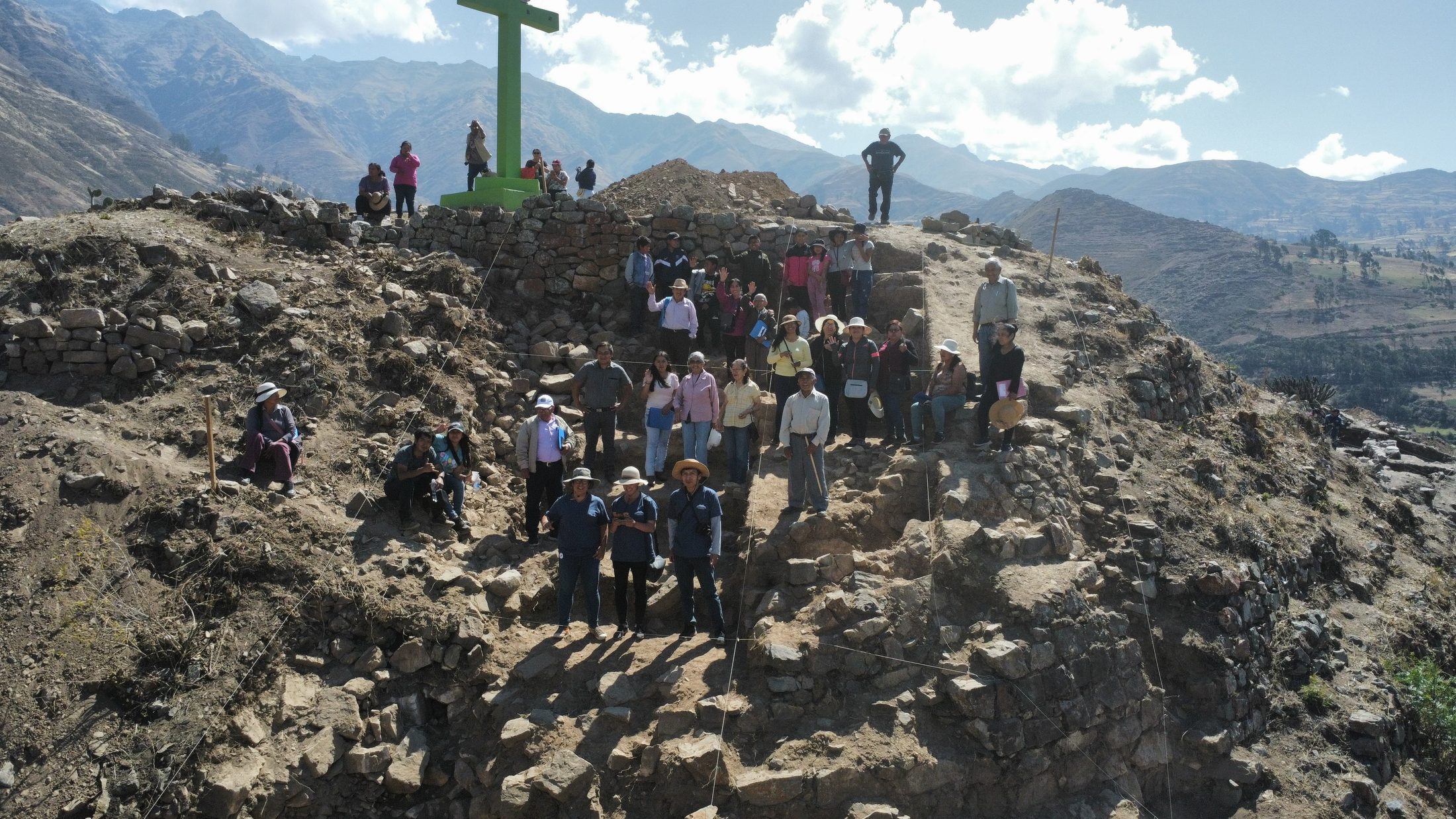 A group of approximately 30 people standing on a hilltop archaeological excavation site with dry rocky terrain, ancient stone walls, and a large green cross, set against a backdrop of Andean mountains and a partly cloudy sky.