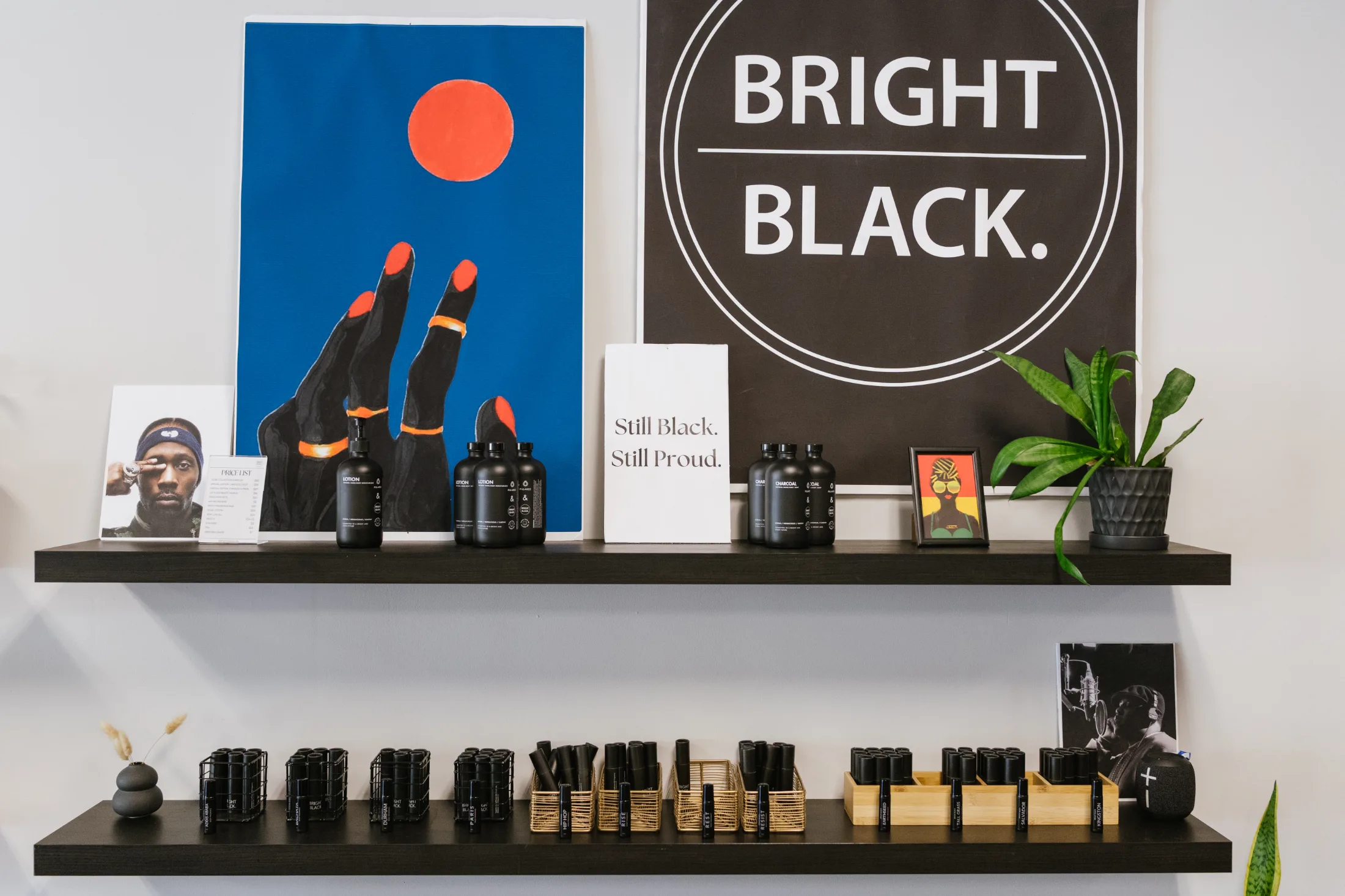 Two black floating shelves display fragrance products, artwork, and plants inside Bright Black’s Durham shop. A large “BRIGHT BLACK.” sign hangs beside a colorful painting of a raised Black hand against a blue background, while a smaller sign reads “Still Black. Still Proud.”